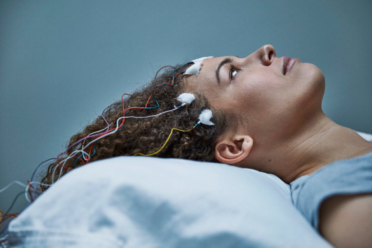 Promotional image for Unrest - Jen Brea, laying on a bed with EEG leads attached to her face and scalp.